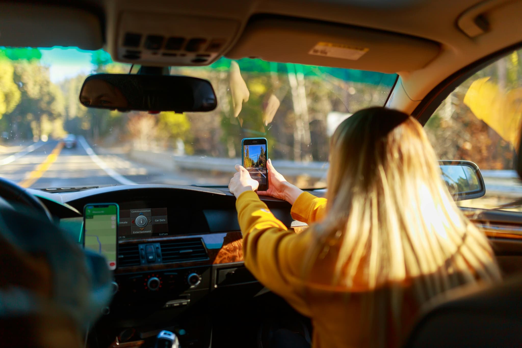 A couple driving in a car, the passenger taking a photo on her phone out of the windscreen.